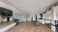 Located at 465 Memorial Dr SE in Atlanta, Georgia, this fitness center offers a thoughtfully designed workout space across a single open floor. The room features warm hardwood flooring that runs throughout, lending a clean and inviting aesthetic to the space. Large black-framed windows line one side of the room, flooding the interior with natural light and offering views of lush, tree-lined surroundings with colorful flowering landscaping — creating an energizing atmosphere for workouts.

A full-length mirror wall spans one side of the room, enhancing the sense of openness while also supporting functional training. Recessed ceiling lighting supplements the natural light, ensuring the space remains bright throughout the day. The fitness center is well-appointed with a variety of equipment, including cycling bikes, a rowing machine, a dumbbell rack with a tiered weight selection, and an assortment of medicine balls and stability balls stored along the wall.

A wall-mounted flat-screen TV is positioned near the mirror for entertainment or guided workout sessions. The space also includes an open floor area suitable for stretching, yoga, or group exercise. Two entry doors, an exit sign, and clean white and gray walls complete the functional, modern design of this well-maintained fitness facility.