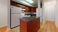 This single-floor unit offers a clean, functional layout with warm hardwood flooring that flows throughout the main living areas. The spacious living room is bright and open, benefiting from a large sliding glass door that brings in generous natural light and provides a view of the surrounding greenery. The space is well-ventilated with visible HVAC vents and maintains a neutral, move-in-ready aesthetic with crisp white walls and clean baseboards.

The kitchen is thoughtfully appointed with rich cherry wood cabinetry, dark granite-style countertops, and a central island that doubles as a prep and dining surface. White appliances — including a refrigerator, electric range, and dishwasher — are neatly integrated alongside a stacked washer and dryer unit, adding everyday convenience. Tile flooring in the kitchen complements the warm wood tones of the adjoining living area.

The bedroom is a comfortable, well-proportioned room with a window fitted with blinds for privacy, and features a reach-in closet with a hanging rod, providing practical storage. The full bathroom includes a soaking tub and shower surrounded by classic white subway tile, tile flooring, and a clean, minimalist finish. Overall, the unit delivers a practical and inviting living environment with cohesive design elements throughout.