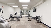Located at 710 N College Ave in Bloomington, Indiana, this fitness center offers a well-organized exercise space designed to accommodate a variety of workout routines. The room features a clean, functional layout with neutral-toned walls, tile-pattern carpet flooring, and bright recessed and flush-mount ceiling lighting that ensures the space feels open and well-illuminated throughout.

The cardio section includes a NordicTrack treadmill, a stationary upright bike, and an elliptical machine, providing options for a range of aerobic workouts. A full-length wall mirror spans one side of the room, visually expanding the space while allowing users to monitor their form during exercise. A wall-mounted flat-screen television adds an entertainment option during cardio sessions.

Strength training is well-supported with a multi-function cable machine suitable for a wide variety of resistance exercises, a dedicated weight bench, and a tiered dumbbell rack stocked with a comprehensive range of free weights. An exercise mat on the floor provides a designated area for stretching or floor-based workouts.

Practical amenities include a paper towel dispenser and waste receptacle near the entrance, supporting cleanliness and hygiene. The space is accessed through a standard door and features an emergency exit sign, indicating it is part of a larger building. Overall, this fitness center is efficiently designed to serve the everyday exercise needs of its users.
