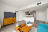 This well-designed 2-bedroom, 2-bathroom residence features a clean, modern aesthetic carried consistently throughout its single-floor layout. The open-concept kitchen and living area serves as the heart of the home, where a peninsula with a white quartz countertop and double-basin sink anchors the space. The kitchen is outfitted with gray two-tone cabinetry, stainless steel appliances including a refrigerator, range, and built-in microwave, complemented by pendant lighting overhead. Wood-look flooring flows through the main living areas, adding warmth to the bright, white-walled interior. The living room is bathed in natural light from a large sliding glass door that opens to a private balcony with open city and palm tree views. Both bedrooms feature plush carpeting and generous windows. The primary bedroom benefits from ample natural light and access to a dedicated closet. Each bathroom is finished with white quartz vanities, modern gray cabinetry with matte black hardware, and wood-look flooring. One bathroom includes a shower-tub combination, while the other features a walk-in shower. The overall ambiance is bright, clean, and contemporary, making efficient use of the available space throughout.