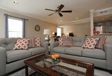 Located in Grapevine, TX, this single-story home offers a comfortable and functional layout designed for everyday living. The main living area is generously sized and anchored by light-toned wood-style flooring that runs throughout, contributing to a bright and airy atmosphere. Recessed lighting in the ceiling is complemented by a ceiling fan, ensuring the space remains well-lit and comfortable year-round. Crown molding adds a clean, finished detail along the upper walls.

The living room is arranged with a plush sectional sofa, accent chairs — including a bold patterned armchair — and a central coffee table, all positioned around a rug-defined seating zone. A wall-mounted flat-screen television serves as a focal point on one side of the room, flanked by a media console and decorative accessories. Natural light filters in through multiple windows dressed with curtains, enhancing the warm, welcoming ambiance.

A dining area with a classic wrought-iron style table and chairs occupies one corner of the space, offering a defined area for meals without sacrificing the openness of the floor plan. The layout flows intuitively, making the home feel cohesive and accessible throughout its single level.