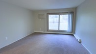 This single-floor space offers a clean, functional layout with a warm and understated aesthetic. The main living area is finished with wall-to-wall carpet in a neutral tone, providing a comfortable and quiet underfoot feel that extends throughout the room. Smooth, light-colored walls create a bright and open atmosphere, complemented by a large sliding glass door that serves as a prominent architectural feature. This expansive glazed opening allows abundant natural light to flood the interior while offering a direct view of the surrounding outdoor greenery. A baseboard heating system runs along the exterior wall beneath the sliding door, ensuring efficient climate control throughout the space. A wall-mounted unit on the adjacent wall provides additional climate management options. The overall ambiance of the space is calm and inviting, with simple, clean lines and a neutral palette that offers a versatile canvas for a variety of interior styles. The single-floor layout provides ease of access and a cohesive flow throughout.