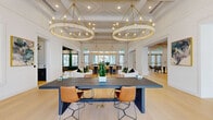 This elegantly designed single-floor clubhouse offers a seamless blend of refined aesthetics and functional communal spaces. The main gathering area commands attention with soaring coffered ceilings adorned with dual oversized brass hoop chandeliers, their warm candlelight glow complementing the pale wide-plank hardwood floors that run throughout. A substantial dark-topped communal table anchors the central space, surrounded by tan leather and wicker seating.

The kitchen area features navy and charcoal cabinetry with brushed gold hardware, dark stone countertops, a large central island with bar seating, and generous windows overlooking the outdoors. Adjacent to the kitchen, a striking emerald-green built-in shelving alcove with marble accents and recessed lighting creates a dramatic focal point, housing a café-style seating nook beneath a coffered ceiling.

A lounge area centers around a floor-to-ceiling dark shiplap fireplace with green herringbone tile surround, paired with plush emerald velvet seating. A bright, airy sitting room features tall windows with sheer drapery and gold curtain rods, flooding the space with natural light. Throughout, white walls, exposed wooden beams, and French doors leading outdoors reinforce the space's bright, coastal-inspired character.