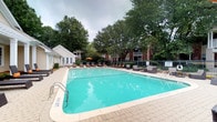 This outdoor amenity space centers around a generously sized swimming pool with clear turquoise water, framed by a wide paved pool deck that provides ample room for movement and leisure. The surrounding area is furnished with rows of cushioned lounge chairs and modular seating arrangements, offering comfortable spots for sunbathing and socializing. A charming single-story clubhouse or amenity structure with white siding and large windows sits adjacent to the pool, providing a convenient gathering or relaxation space. The pool area is enclosed by a wrought iron fence, ensuring privacy and security. Mature, towering trees envelop the space, creating a natural canopy that provides shade and a lush, park-like atmosphere. The backdrop reveals well-maintained brick residential buildings, integrating the pool area seamlessly into the broader community. Colorful accent umbrellas add a vibrant touch to the setting. The combination of thoughtfully arranged outdoor furniture, verdant surroundings, and a well-maintained pool makes this space a welcoming retreat designed to serve as a central hub for outdoor recreation and community gathering.