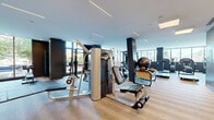 Located at 915 Thayer Ave in Silver Spring, Maryland, this fitness center offers a generously sized, single-floor space designed to accommodate a full spectrum of workout routines. The interior is defined by a contemporary aesthetic, featuring light wood-tone flooring, clean white ceilings with linear LED lighting strips, and dark structural columns that provide bold visual contrast throughout.

Floor-to-ceiling windows line multiple walls, flooding the space with natural light and offering views of the surrounding neighborhood's greenery. Glass doors open directly onto an outdoor terrace, seamlessly connecting indoor training areas to the exterior.

The facility is thoroughly equipped with a wide variety of fitness equipment. A dedicated cardio zone houses a row of treadmills, cycling bikes, and elliptical machines. A strength training area features cable and weight machines, a power rack with Olympic barbells, adjustable benches, and a full dumbbell rack. Interlocking mat flooring in select zones designates functional training and free-weight areas. Large wall mirrors enhance the sense of space and allow users to monitor their form.

A separate enclosed room with hardwood-style flooring and mirrored walls serves as a dedicated cycling studio with additional spin bikes. The overall ambiance is professional, bright, and well-organized, creating an inviting and high-performing fitness environment.