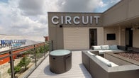 The Party Deck at Circuit is a rooftop amenity space designed to serve as a premier venue for social gatherings and events. Accessed through a dedicated entry door clearly marked "Party Deck," the space transitions from a clean interior corridor—finished with neutral walls, recessed lighting, and dark-framed doors—out onto an open-air deck.

The outdoor deck features a composite wood-style deck surface and is enclosed by sleek glass-panel railings that preserve unobstructed views of the surrounding cityscape and tree canopy. The deck is furnished with woven rattan lounge seating accented by soft teal cushions, along with a coordinating round ottoman-style table, creating a comfortable and stylish setting for entertaining under open skies.

The building's bold "CIRCUIT" signage is prominently displayed on the exterior wall facing the deck, reinforcing the property's brand identity. The interior approach to the deck includes a well-lit transitional hallway with metal stair railings and recessed ceiling lights, providing a polished and functional passage to the outdoor space. Together, the interior and exterior elements create an inviting, versatile amenity suited for both casual socializing and organized events.
