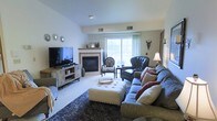 This single-floor residence offers a thoughtfully designed and warmly decorated living space with a distinct eclectic charm. The main living area is the focal point of the home, centered around a classic fireplace with a light wood mantel that serves as a natural gathering point. A large sliding glass door floods the room with natural light, creating a bright and inviting atmosphere while offering a view to the outdoors.

The living room is generously proportioned, accommodating a plush sofa adorned with layered throw pillows in warm tones, a tufted ottoman serving as a central coffee table, and a pair of accent chairs — one upholstered in a bold animal print and another in rich dark leather — that add visual interest and personality to the space. A large flat-screen television is mounted on a rustic media console, balancing entertainment functionality with stylish design.

Decorative wall accents, including a sunburst mirror arrangement and a statement wall sconce, contribute to the room's curated aesthetic. Soft carpeting and a dark area rug add texture and comfort underfoot. The overall ambiance is cozy yet stylish, blending traditional furnishings with eclectic accents to create a lived-in, welcoming environment ideal for both relaxing and entertaining.