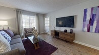 This single-floor residence is thoughtfully designed with a cohesive, contemporary aesthetic carried throughout each room. The living area serves as the heart of the space, featuring wood-look flooring, neutral gray walls, and abundant natural light from multiple windows dressed with grommet-top curtains. A wall-mounted flat-screen television anchors the room above a mid-century modern media console, while a glass-top coffee table and plush seating arranged over a deep plum area rug complete the inviting setup.

The kitchen is well-equipped and stylish, showcasing white shaker-style cabinetry, granite countertops, a stainless steel sink, and a full suite of stainless steel appliances including a gas range, microwave, and refrigerator. A tile backsplash adds texture, and a peninsula with seating provides a casual dining option.

The bedroom offers a restful retreat, highlighted by a bold purple accent wall, soft carpeting, and generous natural light. A walk-through leads to additional storage. The bathroom continues the refined finishes with a granite-topped vanity, white cabinetry, wood-look tile flooring, and a full tub-shower combination. Textured ceilings are present throughout, adding a classic residential character to the overall space.