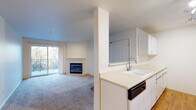 Located at 14700 SW Beard Rd in Beaverton, Oregon, this single-floor unit offers a well-designed, open layout that maximizes functionality and comfort throughout. The living area is generously proportioned, anchored by a classic tile-surround fireplace that serves as a focal point of the room. Soft wall-to-wall carpeting adds warmth underfoot, while a sliding glass door opens directly onto a private balcony overlooking lush, mature trees — bringing the natural beauty of the Pacific Northwest indoors. The kitchen is efficiently laid out with ample white cabinetry, light countertops, wood-look flooring, and a full suite of appliances including a refrigerator, electric range, built-in microwave, and dishwasher. A peninsula counter connects the kitchen to the living space, creating a seamless, open-concept flow. The bedrooms are comfortably sized with plush carpet and large windows that frame verdant outdoor views, filling each room with natural light. One bedroom provides convenient access to the in-unit laundry closet, which houses a full-size washer and dryer. The bathroom features a tub and shower combination with sliding glass doors, a wide vanity, and hardwood-style flooring. Clean, neutral tones throughout lend the space a bright and inviting ambiance.
