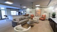 This single-floor communal lounge is thoughtfully designed to encourage gathering and conversation. The main seating area is generously sized, anchored by a plush gray sofa and complemented by a mix of upholstered armchairs in warm coral and neutral tones, arranged around a sleek oval coffee table with a metal base. The layout promotes an open, inviting atmosphere that accommodates both small group interactions and larger social gatherings.

Overhead, multiple ceiling fans with brushed metal finishes provide both functional airflow and a subtle industrial-chic design element. Recessed lighting and flush-mount ceiling fixtures ensure the space is well-lit throughout. The neutral carpeted flooring ties the room together while adding warmth underfoot.

A standout decorative feature is a large framed wall art piece bearing the phrase, "There are no strangers here, only friends you haven't yet met," reinforcing the welcoming, community-oriented character of the space. In the background, a kitchenette with a bar-style counter and bar stools provides a casual spot for light refreshments. A tall floor lamp adds a residential touch, rounding out the approachable, social ambiance of this well-appointed communal area.
