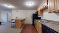 Located at 1010 S Dunn Ave in Fayetteville, Arkansas, this single-floor unit offers a functional and straightforward layout well-suited for everyday living. The kitchen is a standout feature, equipped with light maple cabinetry, granite-style countertops, and a full suite of black appliances including a refrigerator, range, microwave, and dishwasher. Rich hardwood-style flooring runs throughout the kitchen, which also includes a peninsula with bar seating, creating a natural connection to the adjacent living area. The living room is generously sized with neutral carpet, white walls, and windows fitted with blinds that allow for natural light while maintaining privacy. Two bedrooms offer carpeted floors, ample space for furniture, and access to natural light through windowed walls. Each bathroom features a tub and shower combo unit, tile flooring in a stone-like pattern, and light wood vanity cabinetry. The overall design is clean and practical, with a neutral color palette and consistent finishes throughout that create a cohesive, comfortable living environment.