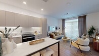 This well-designed 2-bedroom, 1-bathroom residence offers a thoughtfully arranged single-floor layout that maximizes every inch of its compact footprint. The open-plan kitchen and living area form the heart of the home, where flat-panel, greige cabinetry extends floor to ceiling, providing ample storage alongside a clean white countertop, stainless steel appliances, and under-cabinet lighting. A sleek dining table with bar seating bridges the kitchen and living zones seamlessly. The living area is bright and airy, benefiting from large floor-to-ceiling sliding doors that flood the space with natural light and offer outdoor views. Light wood-tone flooring runs continuously throughout, lending warmth and visual cohesion. The primary bedroom is a comfortable retreat, featuring a generous walk-in closet with built-in shelving and hanging rails, a large window dressed with linen drapes, and ample room for a full bedroom suite. The second bedroom is equally bright, with a window framing outdoor views. The bathroom is finished in a clean, contemporary style with white square tile, a soaking tub with shower, and brushed gold fixtures that add a refined touch. Elevated ceilings throughout enhance the overall sense of openness in this modern residence.
