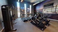 This modern fitness center is thoughtfully designed to support a well-rounded workout experience. The space is anchored by a row of treadmills and elliptical machines positioned along the main floor, all set atop durable, textured carpet tiles. Tall, narrow vertical windows line one wall, flooding the room with natural light and offering views of the surrounding outdoor landscape. Multiple wall-mounted flat-screen televisions are positioned throughout, allowing users to stay entertained during cardio sessions.

A standout design feature is the dramatic large-scale Prada Marfa mural that spans an entire accent wall, adding a bold artistic element to the space and giving it a distinctive, contemporary character. Exposed ductwork runs along the ceiling, complementing the industrial-modern aesthetic, while pendant lighting adds a polished finish overhead.

The free weight area includes a full rack of dumbbells and an adjustable bench, catering to strength training needs. A multi-function cable machine provides additional resistance training options. A water bottle filling station and water fountain are conveniently located along the wall.

A full glass partition wall opens toward an adjacent lounge area, creating a visual connection between the fitness center and the broader amenity space and contributing to an open, inviting atmosphere.