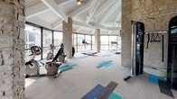 This single-level fitness center is a standout facility defined by its striking architectural character and thoughtful design. Soaring vaulted ceilings with exposed white-painted rafters and tongue-and-groove sheathing create an airy, open atmosphere throughout the space. Prominent natural stone columns — ranging from square to rounded forms — serve as bold structural and aesthetic anchors, lending a warm, rustic contrast to the otherwise contemporary interior.

Floor-to-ceiling windows and glass walls wrap much of the perimeter, flooding the space with abundant natural light and providing sweeping outdoor views, including glimpses of a pool area and surrounding greenery. The flooring is a neutral carpeted surface accented by a vibrant geometric tile pattern in shades of teal, blue, and gray, adding a modern, energetic touch underfoot.

The fitness area is generously proportioned, accommodating a variety of cardio equipment including treadmills, ellipticals, and a recumbent bike, as well as a cable strength-training machine. A dedicated entry zone features a bold teal-and-blue decorative wall installation alongside open blue cubbies for storage, creating a welcoming transition into the workout space. Exposed HVAC ductwork and pendant light fixtures with aqua glass shades complete the interior, balancing industrial and coastal design sensibilities throughout.