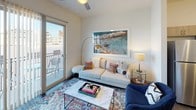 This single-floor residence offers a well-designed open-concept layout with contemporary finishes throughout. The kitchen serves as a central highlight, featuring a large island with a quartz countertop, warm wood-toned cabinetry with brushed metal hardware, stainless steel appliances, and a classic subway tile backsplash. Pendant lighting above the island adds a polished touch, while a dedicated pantry provides ample storage. Dark hardwood-style flooring runs throughout, adding warmth and continuity between spaces.

The living area is bright and inviting, furnished with a comfortable seating arrangement and accented by decorative artwork. A sliding glass door opens directly onto a private balcony, filling the room with natural light and offering outdoor access. A ceiling fan adds both function and style.

The bedroom is a calm, well-proportioned retreat with a large window dressed in layered curtains and blinds, allowing for flexible natural light. Flanked by matching nightstands and lamps, the room provides a serene atmosphere.

The bathroom features wood-look tile flooring, a tub-shower combination with white subway tile surround, open shelving for storage, and clean, neutral finishes that maintain the unit's modern aesthetic throughout.