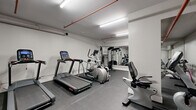 Located at 1161-1171 Wellington St W in Ottawa's vibrant Hintonburg neighbourhood, this fitness room delivers a practical and well-organized workout facility. The single-floor space features clean white walls, durable rubber tile flooring, and bright linear fluorescent lighting that ensures the room remains well-lit throughout. Exposed red pipe runs along the ceiling, adding an industrial character to the otherwise minimalist design.

The cardio area is equipped with a treadmill, an elliptical trainer, and a recumbent exercise bike, providing a range of low- and high-impact options for users. Strength training is supported by a multi-function cable machine, a leg press/extension machine, and an adjustable weight bench, catering to both resistance and functional training needs. A large floor-to-ceiling mirror spans one wall, visually expanding the space and allowing users to monitor their form during exercise.

Climate comfort is maintained by a wall-mounted mini-split air conditioning unit. Safety is addressed with a clearly visible fire extinguisher, and general amenities include a paper towel dispenser and a stainless steel waste bin. The room is thoughtfully arranged to maximize usable floor space, making it an efficient and accessible fitness facility for residents of the building.