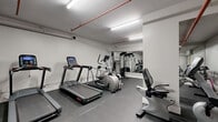 Located at 1161-1171 Wellington St W in Ottawa's vibrant Hintonburg neighbourhood, this fitness room delivers a practical and well-organized workout facility. The single-floor space features clean white walls, durable rubber tile flooring, and bright linear fluorescent lighting that ensures the room remains well-lit throughout. Exposed red pipe runs along the ceiling, adding an industrial character to the otherwise minimalist design.

The cardio area is equipped with a treadmill, an elliptical trainer, and a recumbent exercise bike, providing a range of low- and high-impact options for users. Strength training is supported by a multi-function cable machine, a leg press/extension machine, and an adjustable weight bench, catering to both resistance and functional training needs. A large floor-to-ceiling mirror spans one wall, visually expanding the space and allowing users to monitor their form during exercise.

Climate comfort is maintained by a wall-mounted mini-split air conditioning unit. Safety is addressed with a clearly visible fire extinguisher, and general amenities include a paper towel dispenser and a stainless steel waste bin. The room is thoughtfully arranged to maximize usable floor space, making it an efficient and accessible fitness facility for residents of the building.