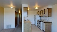 This single-floor space offers a practical and well-organized layout designed for comfortable everyday living. The kitchen serves as a central feature, equipped with a full suite of white appliances including a refrigerator, electric range with oven, and an overhead range hood. Light wood cabinetry lines both the upper and lower sections of the kitchen, providing generous storage space, while a countertop with an integrated sink adds functionality. The warm-toned countertops and light wood finishes lend the kitchen a clean, approachable aesthetic. Ceiling-mounted globe light fixtures provide consistent ambient lighting throughout the space. A structural column with additional cabinetry divides the kitchen from the adjoining hallway, which features carpeted flooring and leads to what appears to be additional rooms and an entry door finished in rich, dark wood. The kitchen itself transitions to light-toned hard flooring, creating a clear distinction between zones. The overall layout is efficient and straightforward, making good use of the available space while maintaining an open, uncluttered feel. The neutral white walls and consistent lighting contribute to a bright, functional ambiance throughout.