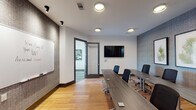 This single-floor professional workspace is thoughtfully designed to accommodate a variety of meeting and collaboration needs. The main conference room serves as the centerpiece, featuring a long rectangular table lined with ergonomic rolling chairs, warm hardwood flooring, and a wall-mounted flat-screen display ideal for presentations. Textured accent walls in neutral tones of gray and brown add visual depth, while recessed and track lighting provide consistent, well-distributed illumination throughout. A large whiteboard mounted on one wall supports brainstorming sessions, and a glass-paneled door connects the space to adjacent areas, maintaining an open, transparent feel.

A smaller breakout room offers a more intimate setting, furnished with a compact table and several chairs beneath framed botanical artwork. Natural light filters in through window blinds, lending a calm, focused atmosphere suited for smaller meetings or private work sessions.

A separate lounge-style meeting area features a round table with upholstered chairs, set beneath a striking pendant light fixture. Floor-to-ceiling windows with horizontal blinds offer views of an outdoor terrace and the surrounding cityscape, bringing in abundant natural light and a sense of openness. Bold abstract artwork completes the room's refined aesthetic.

Together, these spaces create a cohesive, modern professional environment designed for both productivity and comfort.