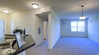 This single-floor residential space offers a clean and functional layout designed for comfortable everyday living. The open-concept floor plan seamlessly connects the kitchen and living areas, creating a cohesive and spacious feel throughout. The kitchen features a dark countertop surface with room for a small bar setup, providing a practical and stylish transition into the main living space. Plush wall-to-wall carpeting extends throughout the living area, adding warmth and comfort underfoot. A large window allows natural light to fill the room, brightening the neutral-toned walls and contributing to an airy ambiance. Architectural details include a partial soffit above the kitchen area, defining the space without enclosing it. A utility panel is neatly integrated into the wall near the hallway corridor, which leads to additional rooms and a doorway visible in the background. The overall color palette is light and neutral, with white baseboards and walls creating a clean, inviting aesthetic. The layout is efficient and well-organized, making the most of the available space while maintaining an open and welcoming atmosphere throughout.