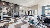 Located at 180 Woodruff Rd in Greenville, SC, this fitness center is a thoughtfully designed, single-floor facility divided into two distinct exercise rooms. The main workout area is a spacious, open room featuring soaring ceilings and a bold teal accent wall that creates an energizing atmosphere. Large windows line the walls, flooding the space with natural light while offering views of the surrounding greenery. A geometric, multi-tone tile floor in shades of white, gray, and brown adds a modern aesthetic throughout. This primary room is outfitted with a comprehensive selection of cardio equipment — including treadmills, ellipticals, a stair climber, and a rowing machine — as well as strength training machines and a full dumbbell rack. Ceiling fans and wall-mounted televisions enhance comfort during workouts. A dramatic floor-to-ceiling athletic mural serves as a focal point on one wall, adding visual energy to the space. The second exercise room offers a quieter, studio-style environment with light wood-look flooring, soft blue walls, and generous window views. This room is equipped with stability balls, yoga mats, and a wall-mounted TV, making it ideal for stretching, yoga, or guided workouts. A large interior window connects the two rooms, maintaining an open and cohesive feel throughout the facility.