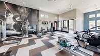 Located at 180 Woodruff Rd in Greenville, SC, this fitness center is a thoughtfully designed, single-floor facility divided into two distinct exercise rooms. The main workout area is a spacious, open room featuring soaring ceilings and a bold teal accent wall that creates an energizing atmosphere. Large windows line the walls, flooding the space with natural light while offering views of the surrounding greenery. A geometric, multi-tone tile floor in shades of white, gray, and brown adds a modern aesthetic throughout. This primary room is outfitted with a comprehensive selection of cardio equipment — including treadmills, ellipticals, a stair climber, and a rowing machine — as well as strength training machines and a full dumbbell rack. Ceiling fans and wall-mounted televisions enhance comfort during workouts. A dramatic floor-to-ceiling athletic mural serves as a focal point on one wall, adding visual energy to the space. The second exercise room offers a quieter, studio-style environment with light wood-look flooring, soft blue walls, and generous window views. This room is equipped with stability balls, yoga mats, and a wall-mounted TV, making it ideal for stretching, yoga, or guided workouts. A large interior window connects the two rooms, maintaining an open and cohesive feel throughout the facility.