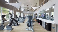 This well-appointed fitness center combines functionality with striking architectural design. The space commands attention with its soaring double-height ceilings, which are adorned with dramatic white sculptural fabric installations that add an artistic flair to the athletic environment. Large windows along the perimeter flood the interior with natural light, creating an energizing atmosphere for workouts.

The layout is thoughtfully organized, with a dedicated cardio zone featuring a long row of treadmills, ellipticals, and stair climbers positioned to take advantage of the natural light and open sightlines. Toward the center of the floor, strength training machines are strategically placed to allow ample space for movement and safe use of equipment.

Along one wall, a continuous built-in counter with dark storage cabinetry provides a functional surface, complemented by wall-mounted televisions for entertainment during workouts. A large mirror further enhances the sense of space. At the rear of the room, colorful exercise balls and additional equipment are neatly organized, adding a vibrant accent to the otherwise neutral palette of gray tones and light wood-grain flooring. Recessed ceiling lighting supplements the natural light throughout.