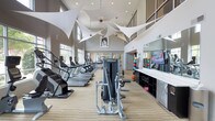 This well-appointed fitness center combines functionality with striking architectural design. The space commands attention with its soaring double-height ceilings, which are adorned with dramatic white sculptural fabric installations that add an artistic flair to the athletic environment. Large windows along the perimeter flood the interior with natural light, creating an energizing atmosphere for workouts.

The layout is thoughtfully organized, with a dedicated cardio zone featuring a long row of treadmills, ellipticals, and stair climbers positioned to take advantage of the natural light and open sightlines. Toward the center of the floor, strength training machines are strategically placed to allow ample space for movement and safe use of equipment.

Along one wall, a continuous built-in counter with dark storage cabinetry provides a functional surface, complemented by wall-mounted televisions for entertainment during workouts. A large mirror further enhances the sense of space. At the rear of the room, colorful exercise balls and additional equipment are neatly organized, adding a vibrant accent to the otherwise neutral palette of gray tones and light wood-grain flooring. Recessed ceiling lighting supplements the natural light throughout.