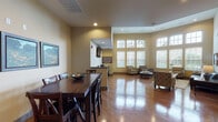 This single-floor communal space is thoughtfully designed to accommodate a variety of activities within one open, well-appointed layout. The most striking architectural feature is the dramatic double-height ceiling in the main lounge area, flanked by expansive floor-to-ceiling windows fitted with horizontal blinds that bathe the interior in generous natural light while framing views of the surrounding greenery outside.

The lounge area is furnished with a dark leather sofa, patterned accent chairs, a glass-topped coffee table, and a round leather ottoman, all arranged atop a neutral area rug over warm hardwood flooring that extends throughout the entire space. Framed landscape artwork adorns the walls, contributing to a polished yet welcoming atmosphere.

Adjacent to the lounge is a dedicated business center alcove, equipped with desk workstations, computers, a printer, and a wall-mounted flat-screen television, making it a functional workspace within the broader communal setting.

The dining area features a dark wood high-top table surrounded by matching cross-back chairs, accompanied by a decorative bowl centerpiece and framed nature prints on the warm-toned walls. A pass-through counter connects to a small service area nearby. Recessed lighting throughout complements the abundant natural light, completing this versatile and inviting shared space.
