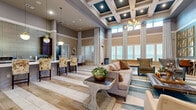 This single-story community clubhouse is designed with both elegance and functionality in mind. The centerpiece is a dramatic main lounge distinguished by soaring double-height ceilings, coffered detailing, and an abundance of natural light pouring through floor-to-ceiling windows dressed with sheer floor-length drapes. Durable wood-look tile flooring runs throughout the common areas, anchoring the warm, neutral color palette accented with teal and gold tones. Comfortable seating arrangements include plush sofas, tufted wingback chairs, and accent tables, creating an inviting atmosphere for socializing or relaxing.

Adjacent to the lounge is a well-appointed kitchenette featuring dark wood cabinetry, a marble-look countertop bar with decorative pendant lighting, stainless steel appliances, and a textured tile backsplash. Eclectic bar-height seating adds a casual dining element.

Private office and meeting areas are furnished with coordinated accent chairs, patterned carpeting, and bold wallcoverings, providing a sophisticated setting for more intimate gatherings or business use.

The fitness center is comprehensively equipped with cardio machines, strength-training equipment, free weights, rubber flooring, large wall mirrors, and natural light from multiple windows—offering a complete workout experience within the facility.