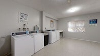 This single-floor communal laundry facility offers a clean, functional layout designed to accommodate shared laundry needs efficiently. The room features multiple top-loading washing machines and front-loading dryers arranged along one wall, providing ample capacity for regular use. A freestanding utility sink is positioned conveniently nearby, offering a space for hand-washing or pre-treating garments, while a sturdy folding table provides a dedicated surface for sorting and folding laundry.

The space is finished with light-toned speckled flooring that is easy to maintain, and the walls are painted in a neutral white-grey palette that keeps the environment bright and clean. Overhead, a flush-mount ceiling light ensures adequate illumination throughout. A window along one wall allows natural light to filter in, contributing to a more open and airy feel despite the compact footprint. A baseboard heating unit runs along the wall, ensuring the room remains comfortable year-round.

Access is provided through a solid navy blue entry door, and informational posted signage for washer and dryer operation is displayed for user guidance. A small fire extinguisher is also present, reflecting attention to safety. Overall, the space is straightforward, well-maintained, and purpose-built for convenient, communal laundry use.