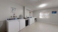 This single-floor communal laundry facility offers a clean, functional layout designed to accommodate shared laundry needs efficiently. The room features multiple top-loading washing machines and front-loading dryers arranged along one wall, providing ample capacity for regular use. A freestanding utility sink is positioned conveniently nearby, offering a space for hand-washing or pre-treating garments, while a sturdy folding table provides a dedicated surface for sorting and folding laundry.

The space is finished with light-toned speckled flooring that is easy to maintain, and the walls are painted in a neutral white-grey palette that keeps the environment bright and clean. Overhead, a flush-mount ceiling light ensures adequate illumination throughout. A window along one wall allows natural light to filter in, contributing to a more open and airy feel despite the compact footprint. A baseboard heating unit runs along the wall, ensuring the room remains comfortable year-round.

Access is provided through a solid navy blue entry door, and informational posted signage for washer and dryer operation is displayed for user guidance. A small fire extinguisher is also present, reflecting attention to safety. Overall, the space is straightforward, well-maintained, and purpose-built for convenient, communal laundry use.