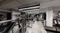 This fitness center is a comprehensively outfitted facility designed to accommodate a wide range of workout preferences. The space is organized into distinct functional zones, each thoughtfully appointed with professional-grade equipment. The main strength training area features a robust selection of cable and resistance machines, adjustable benches, and a full dumbbell rack, all set against a backdrop of large wall-mounted mirrors that enhance the sense of space and allow for proper form monitoring. A flat-screen television is mounted prominently in this zone for entertainment during workouts. The walls throughout are adorned with bold, graphic silhouettes of athletes in motion, reinforcing the energetic ambiance of the facility. Flooring transitions between patterned carpet tiles in the strength zones and rubberized mats in high-traffic equipment areas, providing both durability and comfort. A separate, glass-enclosed cardio room houses spin bikes and upright cycling machines, finished with light hardwood-style flooring and charcoal-toned walls for a focused training environment. The main cardio area includes ellipticals and treadmills. Overhead, linear LED lighting and exposed industrial ceiling elements give the facility a modern, urban character throughout.