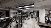 This fitness center is a comprehensively outfitted facility designed to accommodate a wide range of workout preferences. The space is organized into distinct functional zones, each thoughtfully appointed with professional-grade equipment. The main strength training area features a robust selection of cable and resistance machines, adjustable benches, and a full dumbbell rack, all set against a backdrop of large wall-mounted mirrors that enhance the sense of space and allow for proper form monitoring. A flat-screen television is mounted prominently in this zone for entertainment during workouts. The walls throughout are adorned with bold, graphic silhouettes of athletes in motion, reinforcing the energetic ambiance of the facility. Flooring transitions between patterned carpet tiles in the strength zones and rubberized mats in high-traffic equipment areas, providing both durability and comfort. A separate, glass-enclosed cardio room houses spin bikes and upright cycling machines, finished with light hardwood-style flooring and charcoal-toned walls for a focused training environment. The main cardio area includes ellipticals and treadmills. Overhead, linear LED lighting and exposed industrial ceiling elements give the facility a modern, urban character throughout.