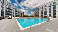 Located at 201 Munson Street in New Haven, Connecticut, this modern multi-story residential complex showcases a striking contemporary architectural design that frames a spectacular central courtyard. The building's exterior blends contrasting materials, including light gray paneling, dark window framing, and bold brick-red accents, creating a visually dynamic facade across multiple floors. Private balconies line the building's interior-facing elevations, offering residents direct views of the vibrant outdoor amenity space below.

At the heart of the complex lies a generously proportioned outdoor swimming pool, finished in vivid aquamarine and surrounded by an expansive stone-tiled deck. The pool deck is well-appointed with rows of white lounge chairs and sun loungers, providing ample space for relaxation. Stylish canopied cabana pods offer shaded retreats for those seeking a more private outdoor experience. Raised planters with greenery add a natural touch to the hardscape, softening the courtyard's modern aesthetic.

The open-air courtyard design allows for abundant natural light, while the multi-story wings of the building create an intimate, resort-like enclosure. The space opens toward the sky, offering sweeping views of the surrounding treetops and New Haven skyline. The overall ambiance is one of upscale, resort-style outdoor living within an urban residential setting.