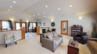 Located at 9295 North Coursier Lane in Spokane, WA, this single-floor property offers an inviting and well-designed layout with a cohesive aesthetic throughout. The interior is defined by soaring vaulted ceilings that lend a sense of openness and airiness to the main living area. Recessed lighting is installed throughout the ceiling, complemented by wall-mounted sconces that provide warm, ambient illumination.

The expansive living room features plush carpeting, generous seating space, and a bold dark accent wall framed by warm wood trim — a detail echoed across doorframes and architectural accents throughout the space. Large windows dressed with wood-slatted blinds flood the room with natural light while offering views of the surrounding outdoor area, including what appears to be a covered patio.

A dedicated media and reading nook is situated within the living area, complete with built-in shelving for books and media storage. The entryway makes a strong first impression with elegant wood-framed French doors that open to the exterior, paired with tile flooring and a nearby dining area featuring a raised table and chairs set beside a window with draping curtains. The overall ambiance is warm, functional, and thoughtfully designed for comfortable everyday living.