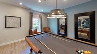 Located at 5100 Quail Run Rd in Riverside, California, this single-floor game and entertainment space is thoughtfully designed for recreation and social gatherings. The layout flows seamlessly from a well-appointed hallway into the main game area, where light hardwood-style flooring, crisp white baseboards, and soft blue-gray walls create a clean, inviting atmosphere throughout.

The centerpiece of the space is a full-size billiards table, positioned beneath a striking multi-tiered drum chandelier that provides both style and focused illumination. A wall-mounted cue rack keeps equipment neatly organized, while a pair of leather accent chairs offers comfortable seating for spectators. Natural light filters in through a curtained window, adding warmth to the room.

Adjacent to the billiards area, a freestanding electronic dartboard cabinet provides an additional entertainment option, accompanied by framed wall art that adds visual depth and personality to the space.

A built-in bar counter with bar-height seating for two serves as a refined gathering spot, accented by elegant pendant lighting and carefully curated artwork along the surrounding walls. Recessed ceiling lighting throughout ensures the entire space is well-lit and functional. The overall design balances a modern aesthetic with a relaxed, recreational ambiance, making it an ideal venue for entertainment and leisure.