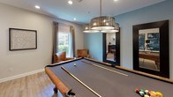 Located at 5100 Quail Run Rd in Riverside, California, this single-floor game and entertainment space is thoughtfully designed for recreation and social gatherings. The layout flows seamlessly from a well-appointed hallway into the main game area, where light hardwood-style flooring, crisp white baseboards, and soft blue-gray walls create a clean, inviting atmosphere throughout.

The centerpiece of the space is a full-size billiards table, positioned beneath a striking multi-tiered drum chandelier that provides both style and focused illumination. A wall-mounted cue rack keeps equipment neatly organized, while a pair of leather accent chairs offers comfortable seating for spectators. Natural light filters in through a curtained window, adding warmth to the room.

Adjacent to the billiards area, a freestanding electronic dartboard cabinet provides an additional entertainment option, accompanied by framed wall art that adds visual depth and personality to the space.

A built-in bar counter with bar-height seating for two serves as a refined gathering spot, accented by elegant pendant lighting and carefully curated artwork along the surrounding walls. Recessed ceiling lighting throughout ensures the entire space is well-lit and functional. The overall design balances a modern aesthetic with a relaxed, recreational ambiance, making it an ideal venue for entertainment and leisure.