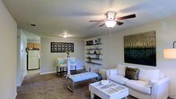 This single-story home offers a thoughtfully arranged living space that balances comfort and style. The main living area is carpeted and generously sized, furnished with a white sofa and a rustic coffee table that lend a casual, inviting feel. A ceiling fan with an integrated light fixture provides both airflow and ambient lighting, while a second flush-mount light ensures the space is well-lit throughout. Built-in open shelving lines one wall, offering both display and storage space for décor and personal items, complemented by a bold piece of wall art featuring a nature-inspired landscape. A decorative lattice-style wall panel adds an artistic touch near the dining nook, where a small high-top table and stools create an intimate spot for casual dining or work. The neutral wall tones and light-colored ceiling contribute to a clean, airy ambiance throughout the living space. A hallway visible in the background suggests additional rooms beyond, completing this practical and comfortably appointed single-floor home.