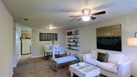 This single-story home offers a thoughtfully arranged living space that balances comfort and style. The main living area is carpeted and generously sized, furnished with a white sofa and a rustic coffee table that lend a casual, inviting feel. A ceiling fan with an integrated light fixture provides both airflow and ambient lighting, while a second flush-mount light ensures the space is well-lit throughout. Built-in open shelving lines one wall, offering both display and storage space for décor and personal items, complemented by a bold piece of wall art featuring a nature-inspired landscape. A decorative lattice-style wall panel adds an artistic touch near the dining nook, where a small high-top table and stools create an intimate spot for casual dining or work. The neutral wall tones and light-colored ceiling contribute to a clean, airy ambiance throughout the living space. A hallway visible in the background suggests additional rooms beyond, completing this practical and comfortably appointed single-floor home.