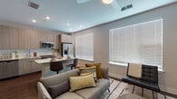 This single-floor unit offers a thoughtfully designed open-concept layout that seamlessly connects the living, dining, and kitchen areas. The living space is anchored by a comfortable sofa accented with gold and earth-toned throw pillows, complemented by a textured area rug, a walnut-finished coffee table, and two large windows fitted with white horizontal blinds that allow for ample natural light. Recessed lighting and a ceiling fan complete the airy feel of the space.

The kitchen is outfitted with a full suite of stainless steel appliances, including a side-by-side refrigerator with an in-door water and ice dispenser, a slide-in electric range, an over-the-range microwave, and a stainless steel sink. Light wood-grain cabinetry and a subtle tile backsplash give the kitchen a clean, modern aesthetic, while a breakfast bar with seating provides a casual dining option.

The bedroom is private and cozy, accessed through a stylish sliding barn door. It features dark hardwood flooring, a storage bed with a woven wood headboard, built-in shelving, and warm ambient lighting.

The bathroom is finished with a wide vanity featuring a white countertop and wood-grain cabinetry, a large framed mirror, recessed lighting, a combination shower and soaking tub with subway tile surround, and dark hardwood-style flooring that ties the space together cohesively.