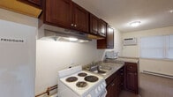 Located at 68 Parker St in Acton, Massachusetts, this single-floor unit offers a practical and straightforward layout designed for comfortable everyday living. The kitchen is a standout feature, outfitted with rich dark-stained wood cabinetry extending to the upper walls for generous storage, granite-style countertops, a double-basin stainless steel sink, and a white electric coil-burner range with an overhead ventilation hood. A wall-mounted air conditioning unit and a window providing natural light complete the kitchen space, which transitions seamlessly into the adjoining living area via tile flooring. The living and bedroom areas are finished with neutral wall-to-wall carpeting and include baseboard heating units for year-round comfort. The bedroom is notably well-appointed with two sliding-door closets offering substantial storage capacity. The full bathroom features tile flooring, a full-size soaking tub with an overhead shower, a toilet, and a dark wood vanity that complements the cabinetry found throughout the unit. The overall layout is compact yet efficient, with a clean, neutral color palette of soft whites and warm beiges that lends a bright and airy ambiance throughout the space.