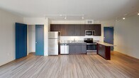 Located at 500 E 14th St in Minneapolis, MN, this modern unit offers a well-designed open-concept layout that seamlessly connects the kitchen and living area. Light-toned hardwood-style flooring flows throughout, complementing the clean white walls and creating a bright, airy atmosphere.

The kitchen is a standout feature, equipped with stainless steel appliances including a refrigerator, range, microwave, and dishwasher. Rich dark-toned cabinetry extends to the ceiling, providing generous storage, while granite countertops with warm tones add a touch of elegance. A breakfast bar offers additional prep and dining space.

The expansive living area is bathed in natural light, courtesy of large black-framed windows that frame impressive views of the Minneapolis skyline and surrounding cityscape. Track lighting runs along the ceiling throughout the space, offering flexible and modern illumination.

The bathroom is tastefully appointed with a dark-toned vanity, a clean white countertop, a mirror with overhead lighting, and a glass-enclosed shower. Bold teal-blue interior doors throughout the unit add a distinctive pop of color and contemporary character.

The unit's single-floor layout ensures ease of navigation, making it both functional and stylish for everyday urban living in the heart of Minneapolis.