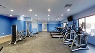 This fitness center offers a thoughtfully designed workout space spread across a single floor. The room is defined by vibrant blue accent walls that contrast with a clean white ceiling, creating an energetic and motivating atmosphere. Recessed lighting throughout the ceiling provides bright, even illumination across the entire space, ensuring excellent visibility during workouts.

The cardio area is prominently featured along one side of the room, lined with a row of treadmills and elliptical machines, all positioned to face a wall-mounted flat-screen television — ideal for staying entertained during extended sessions. On the opposite side, strength training equipment, including a cable machine and weight bench with free weights, provides users with a comprehensive resistance training setup.

The flooring consists of durable, textured carpet designed to withstand heavy equipment and high foot traffic while providing comfort underfoot. A large mirror along one wall enhances the sense of space and allows users to monitor their form. A window along one wall allows natural light to filter in, adding to the open and welcoming feel of the facility.