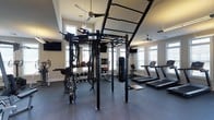 Located on Langdon Road in Morrisville, NC, this fitness center offers a comprehensive workout environment designed to support a wide range of training styles. The space is anchored by a large multi-functional rig system outfitted with monkey bars, battle ropes, a heavy punching bag, and a variety of attachments, making it ideal for functional and strength-based training. A dedicated free weight area features an organized rack of kettlebells and medicine balls, providing options for dynamic and full-body workouts. The cardio zone includes multiple treadmills positioned near large, bright windows that invite abundant natural light throughout the space. Mounted flat-screen televisions are positioned throughout the room for entertainment during workouts. The facility benefits from vaulted ceilings that lend an open, airy feel, further enhanced by ceiling fans that promote air circulation. Durable rubberized flooring covers the workout area, ensuring safety and functionality. The neutral wall tones and ample window placement create a clean, energizing atmosphere well-suited for both focused individual training and group fitness activities.