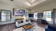 This well-appointed single-floor space functions as a community clubhouse or leasing office, designed with a contemporary aesthetic that balances comfort and professionalism. The main lounge area is expansive and airy, benefiting from soaring ceilings with a central skylight that floods the room with natural light. Rich wood-look flooring runs throughout, complementing a bold two-tone color scheme of crisp white and deep navy blue accent walls that lend the space a polished, modern character.

The primary gathering area is anchored by a large wall-mounted display screen and furnished with a white sofa, patterned throw pillows, and a rustic wooden coffee table set atop a geometric area rug. A glass-top dining table with modern chairs provides a casual seating nook near the black-framed windows, which offer views of the outdoor surroundings.

A secondary lounge area features a dramatic floor-to-ceiling dark marble-tiled accent wall housing a sleek linear fireplace, flanked by a pair of plush royal blue velvet chairs. Glass entry doors and additional windows introduce generous natural light and outdoor views to this seating zone. A dedicated workstation area and a service counter add functional utility to the space. A hallway connects to a restroom, decorated with curated artwork, completing this thoughtfully designed environment.