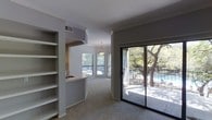 This single-floor residence offers a well-organized layout that balances open living spaces with functional private areas. The main living area is carpeted and bathed in natural light, thanks to multiple windows and a full-width sliding glass door that opens onto a private balcony. The balcony overlooks a pool and mature trees, providing a serene outdoor connection. Crown molding runs along the ceiling, adding a refined architectural detail to the space.

A built-in shelving unit lines one wall of the living area, offering ample storage and display space. The kitchen is separated from the living area by a half-wall with a pass-through counter, maintaining visual openness while defining the cooking space. The kitchen features white cabinetry and a sink positioned near the window, allowing natural light to fill the area.

The bathroom includes essential fixtures including a toilet, sink, and additional cabinetry, finished with dark hardwood-style flooring that contrasts with the lighter tones found elsewhere. The overall color palette throughout the unit is neutral, with white and gray tones creating a clean, cohesive ambiance suited to a variety of interior styles.