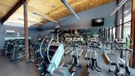 This fitness center offers a comprehensive workout environment housed within a thoughtfully designed single-floor space. The architectural character is immediately established by striking warm wood-paneled ceilings supported by exposed timber beams, which lend a natural, lodge-inspired aesthetic to the facility. Large floor-to-ceiling windows line one side of the space, flooding the interior with natural light and providing sweeping views of lush tropical greenery and outdoor surroundings, creating an energizing atmosphere for exercise.

The cardio zone is generously appointed with a full lineup of treadmills, elliptical trainers, and stair climbers, all arranged in orderly rows. Wall-mounted televisions are positioned for easy viewing during cardio sessions. Full-length mirrors along the walls enhance the sense of openness and allow for proper form monitoring throughout the space.

The strength training area is equally well-stocked, featuring multiple dumbbell racks with a wide range of weights, as well as a variety of selectorized cable and resistance machines suited for both upper and lower body workouts. Rubber flooring throughout provides safety and durability across all zones.

Ceiling fans and linear pendant lighting complement the overall design, ensuring comfort and visibility. The combination of natural materials, abundant daylight, verdant outdoor views, and extensive equipment makes this fitness center a functional and visually inviting space.