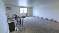 This two-floor space offers a straightforward and functional layout designed for comfortable everyday living. The upper level features a generously sized open living area finished with wall-to-wall carpeting that adds warmth underfoot. A window on the far wall allows natural light to fill the room, creating a bright and airy atmosphere. A baseboard heating system runs along the exterior wall, ensuring efficient climate control throughout the colder months. The staircase connecting the two floors is framed by a sturdy metal railing, providing both safety and a subtle industrial accent to the otherwise neutral interior. The walls are finished in a clean, light tone that enhances the sense of openness throughout the space. The overall design is simple and uncluttered, prioritizing usable square footage and ease of movement. The neutral palette and practical finishes make this space highly adaptable to a variety of furnishing styles and personal preferences. With its open upper-level living area and multi-floor layout, the space offers a versatile and livable environment suited to a range of occupants.