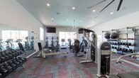 This single-level fitness center offers a comprehensive workout environment designed to accommodate a variety of training styles and fitness goals. The space is defined by its clean, white walls, elevated ceilings, and recessed lighting, all of which contribute to an open and energizing atmosphere. Large windows along the perimeter allow natural light to fill the room, enhancing the sense of space throughout.

The cardio zone is well-appointed with multiple treadmills, elliptical trainers, and a rowing machine, positioned to take advantage of the natural light and outward views. A wall-mounted flat-screen television provides entertainment for cardio sessions. Overhead, a large ceiling fan ensures consistent airflow across the space.

The strength training area includes a multi-station cable machine, a Smith machine, a functional trainer, and a dedicated free weights section featuring a full rack of dumbbells in varying sizes. Additional equipment such as a punching bag, kettlebells, medicine balls, resistance bands, and a weight bench further support functional and strength-based training.

Mirrored walls along one side of the room visually expand the space and assist with form checking during workouts. A water fountain is conveniently accessible, and the patterned carpet flooring adds both durability and visual interest to the overall design.